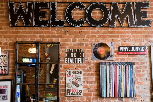 A brick wall with a large "WELCOME" sign, framed mirrors, and various decorative signs and vinyl records.