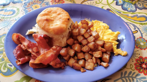 A blue plate with a biscuit, scrambled eggs, crispy bacon, and golden-brown diced potatoes on a colorful tablecloth.