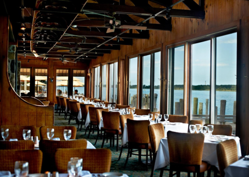A cozy restaurant with wooden decor, featuring tables set by large windows overlooking a serene waterfront view.