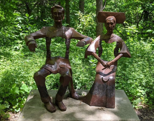 A metal sculpture of a seated man and woman in a lush green forest setting.