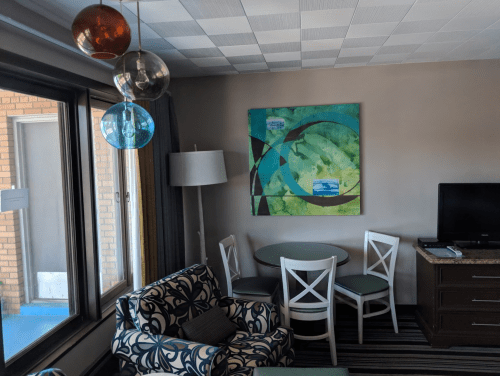 A cozy hotel room featuring a patterned chair, round table, TV, and colorful pendant lights, with a large abstract painting.