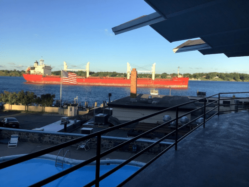 A large cargo ship sails on a river, with a pool and flag in the foreground and a clear blue sky above.