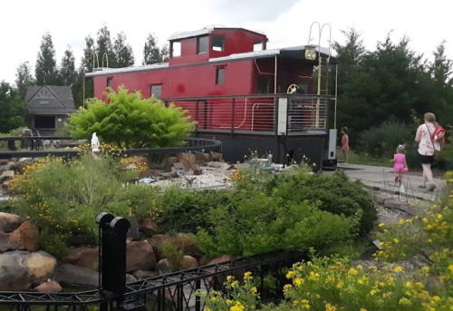 A red train caboose converted into a building, surrounded by greenery and flowers, with people walking nearby.
