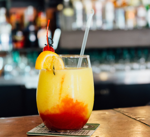 A colorful tropical drink with orange and red layers, garnished with a cherry and an orange slice, on a bar counter.