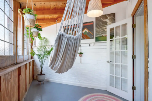 A cozy indoor space featuring a hanging chair, plants, artwork, and large windows with natural light.