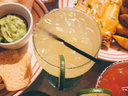 A refreshing margarita with ice and a lime wedge, surrounded by chips, guacamole, and a plate of enchiladas.