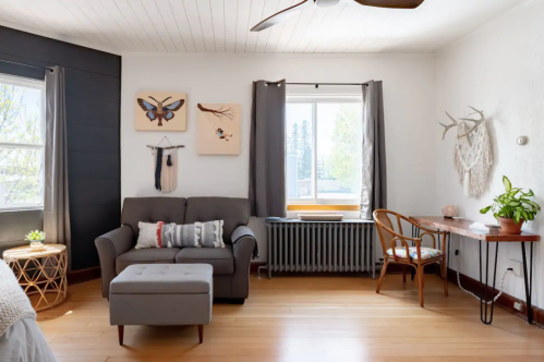 Cozy living room with a gray sofa, wooden desk, plants, and wall art, featuring natural light from a window.