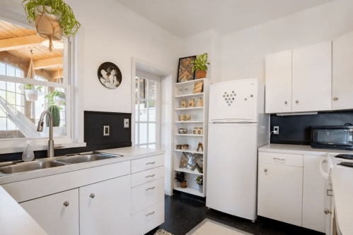 Bright kitchen with white cabinets, a refrigerator, and a shelf of jars, featuring plants and natural light.