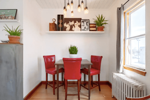 Cozy dining nook with a wooden table, four red chairs, plants, and a shelf displaying framed photos and decor.