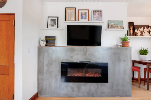 Modern living room with a concrete fireplace, TV above, and shelves displaying framed photos and plants.
