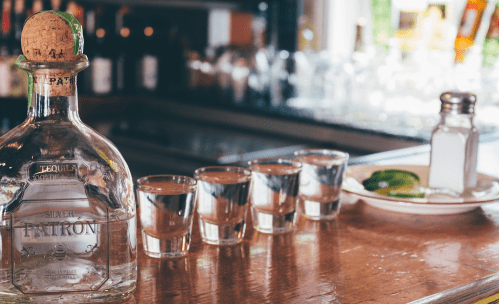 A bottle of Patron tequila with four shot glasses and a plate of lime wedges on a bar counter.