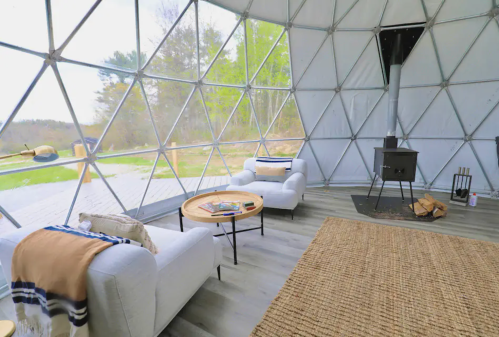 Cozy interior of a geodesic dome with two chairs, a coffee table, and a wood stove, surrounded by greenery outside.