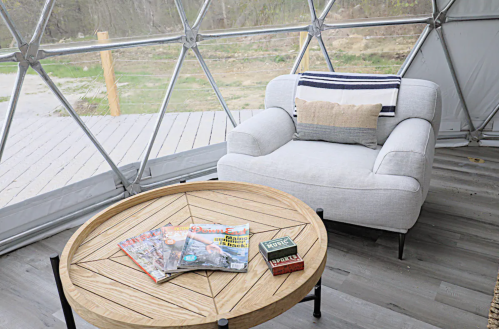 Cozy interior of a geodesic dome with a light gray armchair and a wooden coffee table stacked with magazines.