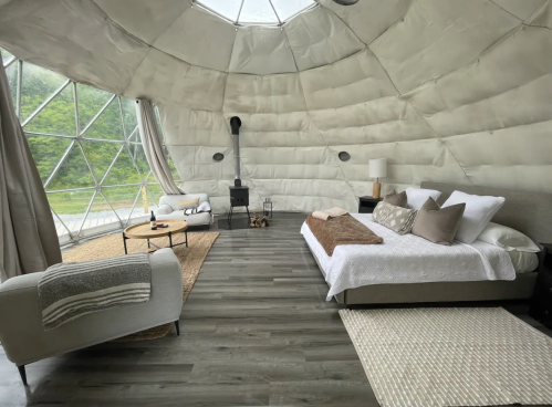 Cozy, modern interior of a geodesic dome with a bed, seating area, and large windows overlooking nature.