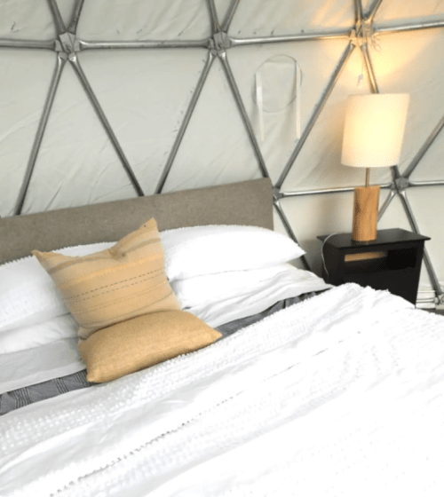Cozy bedroom inside a geodesic dome, featuring a bed with white linens, a decorative pillow, and a wooden lamp.