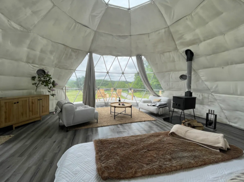 Cozy interior of a geodesic dome with a bed, seating area, and large windows overlooking a green landscape.