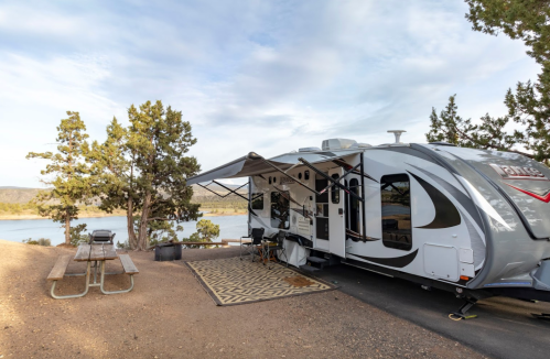 A parked RV by a lake, with a picnic table nearby and trees in the background under a partly cloudy sky.