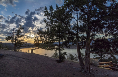 Sunset over a calm lake, framed by trees and a cloudy sky, creating a serene natural landscape.