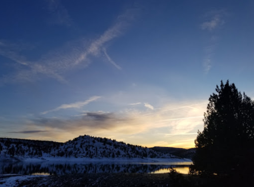 A serene sunset over a snowy landscape, with a calm lake reflecting the colorful sky and silhouetted trees.
