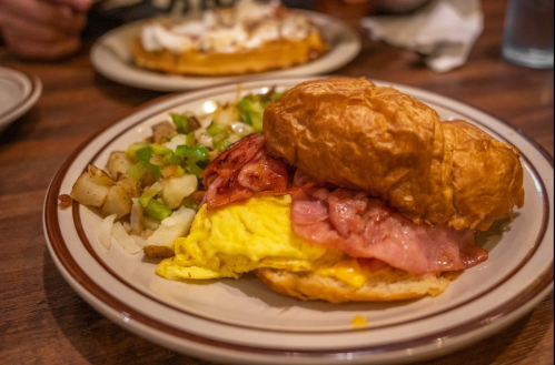 A croissant sandwich filled with ham, egg, and cheese, served with diced potatoes and green onions on the side.