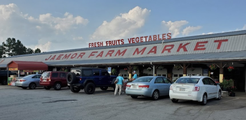 Exterior of Jaemor Farm Market with a sign for fresh fruits and vegetables, featuring parked cars and a blue sky.