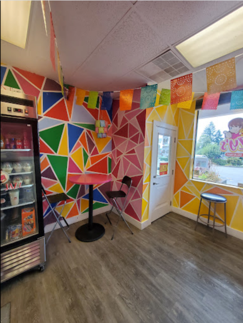 Colorful interior of a cafe with geometric wall art, a small table, chairs, and a fridge. Brightly decorated with banners.