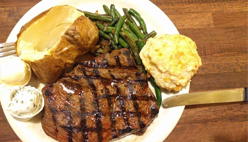 A plate with grilled steak, baked potato, green beans, and a biscuit, accompanied by butter and herb sauce.