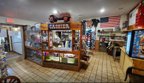 A cozy shop interior featuring a wooden cashier counter, souvenirs, and Route 66 memorabilia, with an American flag on the wall.