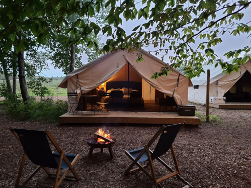 A cozy glamping setup with a fire pit and chairs in front of a tent surrounded by trees.