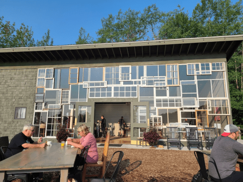 A modern building with a unique facade made of various window frames, surrounded by outdoor seating and greenery.