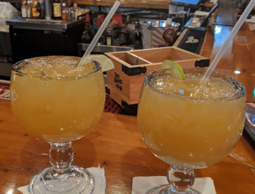 Two large margaritas with lime slices and straws, served on a wooden bar counter.