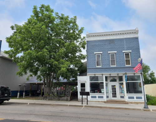 A blue two-story building with large windows, an American flag, and a tree in front, set against a cloudy sky.