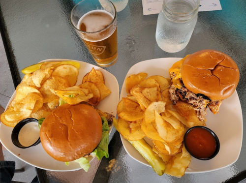 Two plates of food: one with a burger and chips, the other with pulled pork and chips, alongside a glass of beer.
