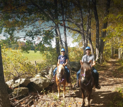 Two people on horseback riding along a wooded trail with autumn foliage.