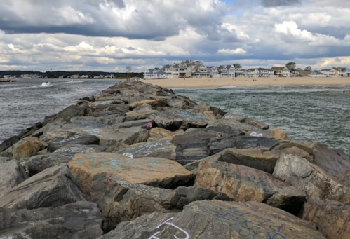 A rocky jetty extends into the water, with a beach and houses visible in the background under a cloudy sky.
