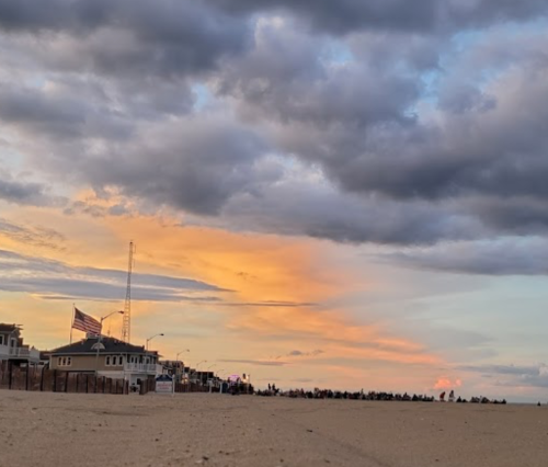 A beach scene at sunset with clouds and a hint of orange in the sky, lined with houses and a distant crowd.