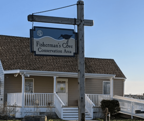 Sign for Fisherman's Cove Conservation Area in front of a light-colored building with a porch. Clear blue sky above.