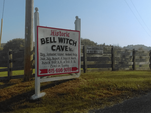 Sign for Bell Witch Cave with visiting hours and contact information, set against a grassy landscape and wooden fence.