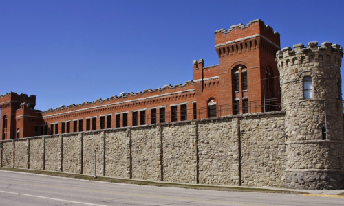 Historic brick building with a stone wall, featuring towers and a clear blue sky in the background.