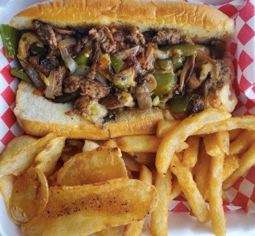 A loaded cheesesteak sandwich with beef, peppers, and mushrooms, served with crispy French fries on the side.