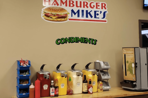 Condiment station at Hamburger Mike's, featuring various sauces and a drink dispenser.