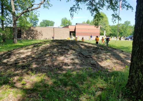 A grassy mound in a park with a group of people in yellow shirts in the background near a building and trees.