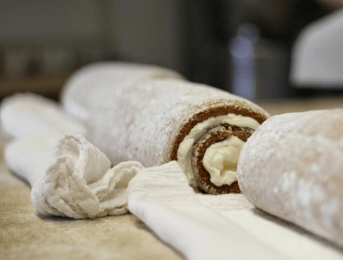 A close-up of a rolled cake dusted with powdered sugar, revealing creamy filling inside, resting on a cloth.