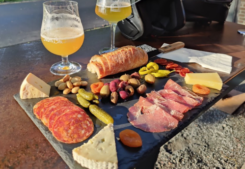 A slate platter with assorted meats, cheeses, olives, pickles, nuts, and a loaf of bread, accompanied by two glasses of beer.