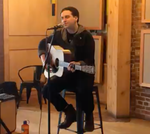 A musician sits on a stool, playing an acoustic guitar and singing into a microphone in a cozy, wooden space.