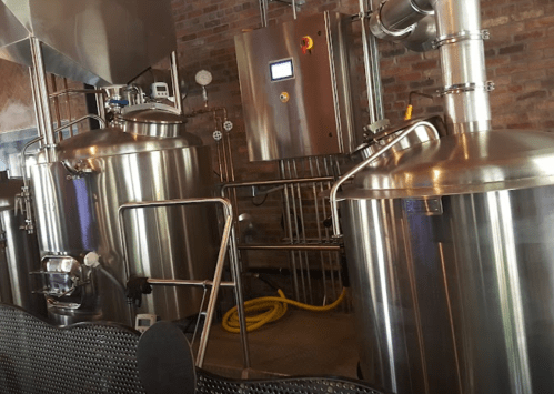 A brewery interior featuring large stainless steel fermentation tanks and brewing equipment against a brick wall.