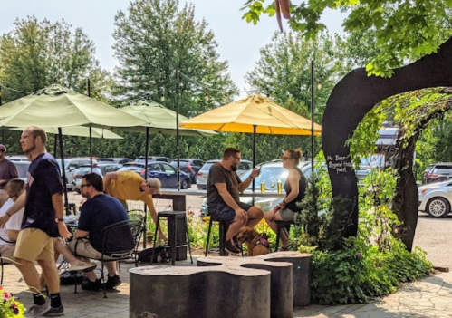 A lively outdoor café scene with people sitting under yellow umbrellas, surrounded by greenery and parked cars in the background.