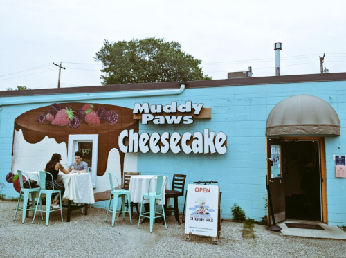 Exterior of Muddy Paws Cheesecake, featuring a colorful sign and outdoor seating with two people at a table.