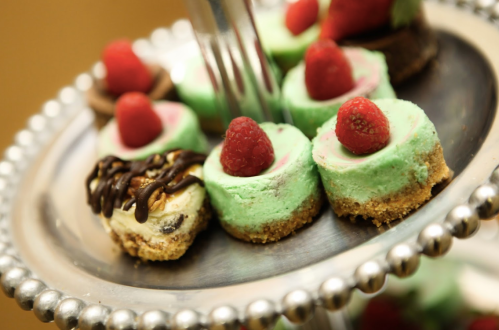 A silver platter holds mini cheesecakes topped with raspberries, featuring green and chocolate layers.