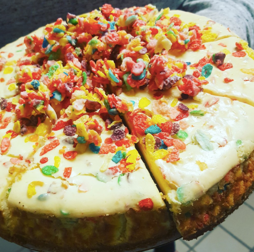 A colorful cake topped with a creamy layer and sprinkled with vibrant cereal pieces, partially sliced.